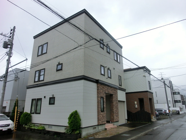 白とクリーム系の外壁に窓枠などにこげ茶のアクセントカラーがシックな佇まいの３階建て戸建て住宅